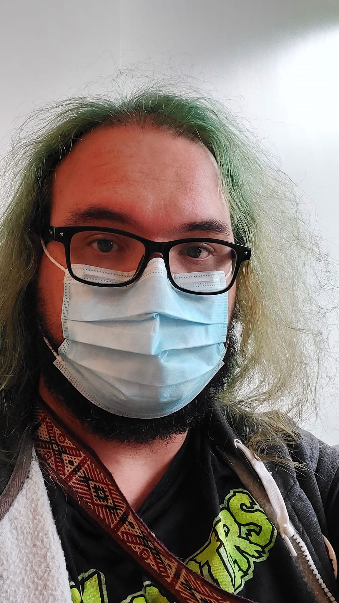 A man with long green hair and a surgical mask stands in a hospital waiting room with white walls